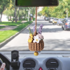 May include: A brown crocheted car air freshener with a basket of colourful flowers, including yellow, white, and purple blooms. The charm hangs from a brown cord and has a small yellow ball at the bottom, adding a handmade touch to any vehicle.