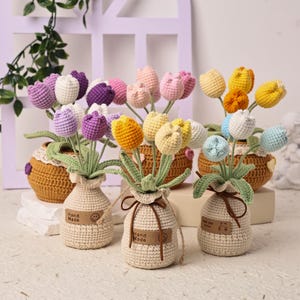 以下が含まれることがあります： 黄色、紫、ピンクなど、さまざまな色の手作りのかぎ針編みのチューリップのコレクション。 チューリップは、小さな織りのバスケットとベージュの布の花瓶に配置されています。 各花瓶には「Hand Made」と書かれたタグが付いています。