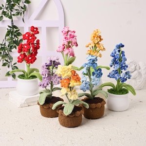 Op de afbeelding: Een verzameling handgemaakte gehaakte bloemen in verschillende kleuren, waaronder rood, roze, geel, blauw en paars. De bloemen staan in kleine witte en bruine potten, met groene bladeren. De opstelling staat tegen een witte achtergrond.