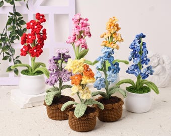 May include: A collection of handmade crochet flowers in various colors, including red, pink, yellow, blue, and purple. The flowers are potted in small white and brown pots, with green leaves. The arrangement is set against a white background.