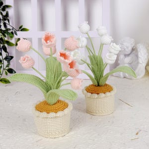 May include: Two crochet tulip plants in cream-coloured pots with scalloped edges. One plant has pink and peach tulips, the other has white tulips. The pots have a yellow-orange base, green leaves, and are set against a white background.