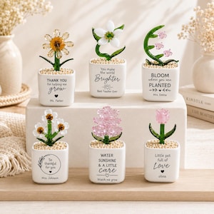 May include: Six small white ceramic square pots, each containing a glass flower sculpture with green stems and small beige pebbles. The pots feature black printed text including phrases like Thank you for helping me grow and Bloom where you are planted.