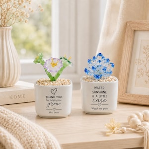 May include: Two white square ceramic planters containing glass succulents. The left planter features a purple and green glass flower and text reading "THANK YOU for helping me grow" with "Mrs. Smith". The right planter has a blue glass succulent and text reading "WATER SUNSHINE & A LITTLE care Watch me grow".