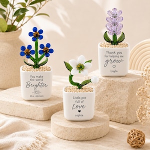 May include: Three small white ceramic pots containing glass flowers. The left pot has blue flowers and reads "You make the world Brighter" with "Mrs. Johnson". The centre pot has a white flower and reads "Little pot full of Love" with "Sophia". The right pot has purple flowers and reads "Thank you for helping me grow" with "Layla".