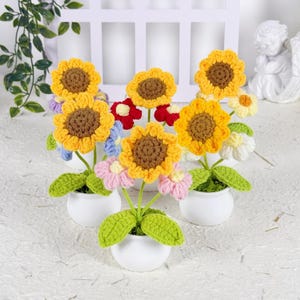 May include: A collection of handmade crochet flowers, including sunflowers, daisies, and other colorful blooms. The flowers are in small white pots with green leaves, creating a cheerful and decorative display.