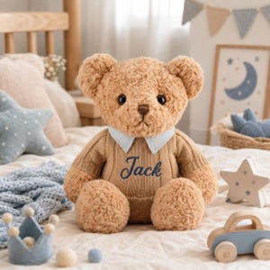 May include: A light brown plush teddy bear with dark eyes and a brown nose. The bear wears a tan ribbed knit sweater with a light blue collar and the name Jack embroidered in dark blue script on the front of the sweater chest area.