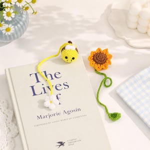 May include: Two handmade crochet bookmarks resting on a book titled The Lives of. One bookmark features a yellow and black bee attached to a white flower by a yellow yarn string. The second bookmark is a brown and orange sunflower with a green stem.