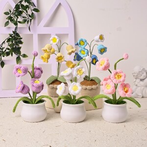 Peut inclure: Plusieurs fleurs crochetées à la main dans des pots en céramique blancs. Les fleurs sont de différentes couleurs, dont le rose, le jaune, le bleu et le violet, avec des tiges et des feuilles vertes. Les fleurs sont dans de petits paniers tressés.