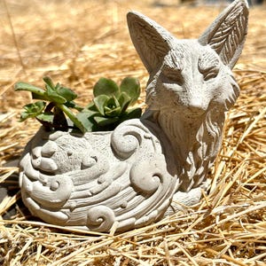 May include: A white concrete fox planter with a detailed design. The planter is filled with green succulents.
