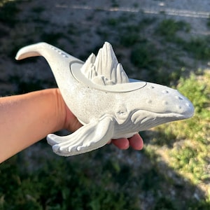 May include: A gray concrete whale sculpture with a mountain range detail on its back. The whale has a curved tail and flippers, and is held by a person's hand. The sculpture is designed to hold small plants or items.