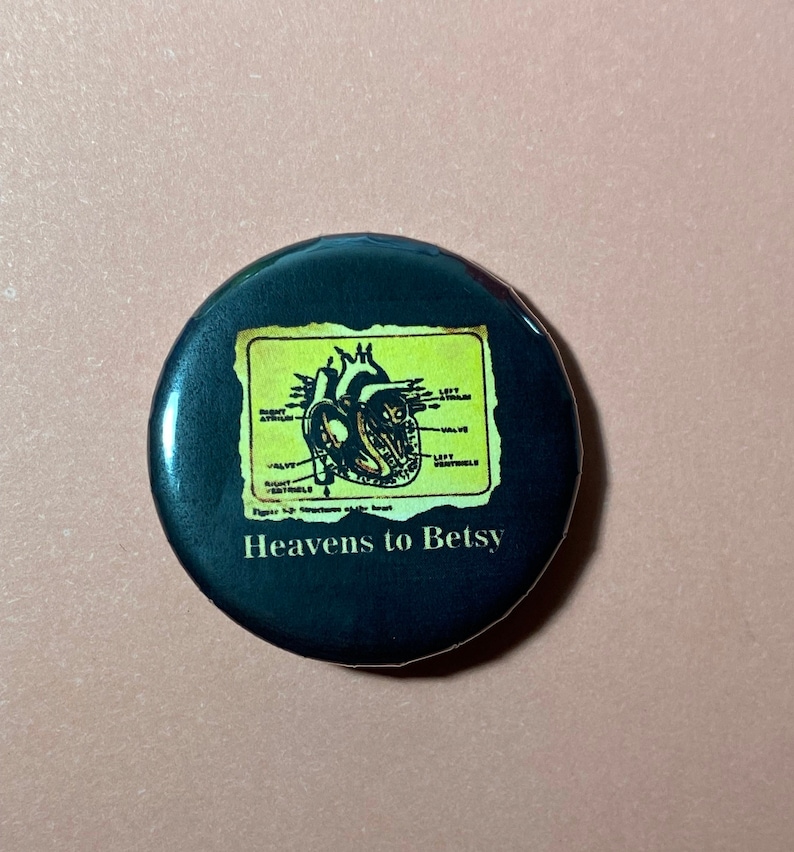 Pu&ograve; includere: Un bottone nero con un'immagine gialla e marrone di un cuore umano con etichette anatomiche. Il testo "Heavens to Betsy" &egrave; stampato sotto l'immagine.