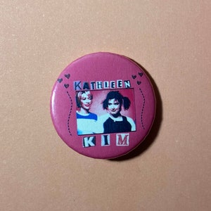 Pu&ograve; includere: Un bottone rosa con una foto di due donne. Il testo "KATHLEEN KIM" &egrave; stampato sul bottone.