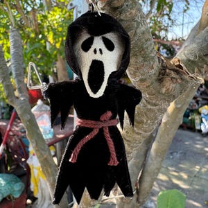 May include: A black and white Scream mask keychain. The keychain features a black hooded robe, a brown rope belt, and a silver metal keychain. The mask has a white face with black eye sockets and a wide open mouth.