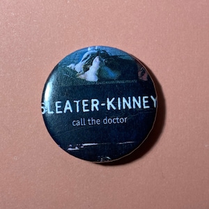 Pu&ograve; includere: Un bottone bianco e nero con il nome della band "Sleater-Kinney" e il testo "call the doctor" stampato su di esso.