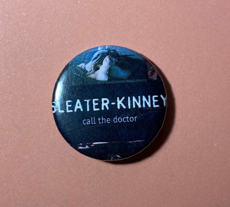 Pu&ograve; includere: Un bottone bianco e nero con il nome della band "Sleater-Kinney" e il testo "call the doctor" stampato su di esso.