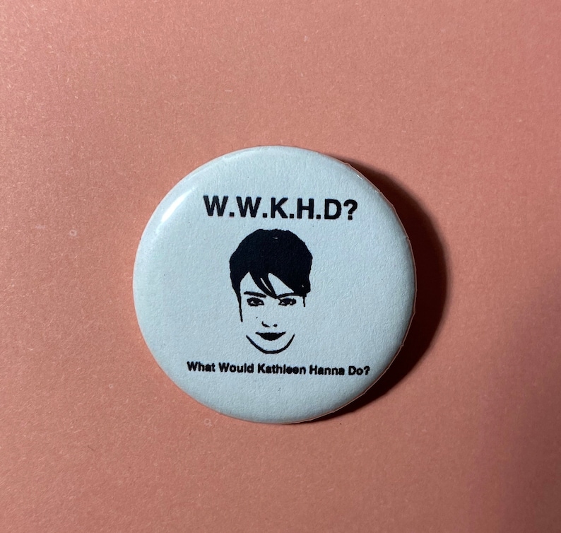Pu&ograve; includere: Bottone bianco con testo nero e un'illustrazione in bianco e nero del viso di una donna. Il testo dice "W.W.K.H.D? What Would Kathleen Hanna Do?"