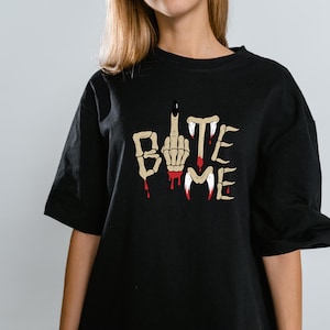Pode incluir: T-shirt preto com a frase "BITE ME" em uma fonte estilizada. O design inclui uma mão com o dedo médio levantado e detalhes de sangue pingando. A camisa é um estilo casual de manga curta.