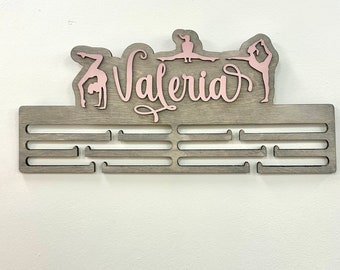 May include: A wooden medal display with the name "Valeria" in pink lettering. The display has eight slots for medals and features three pink gymnast silhouettes.