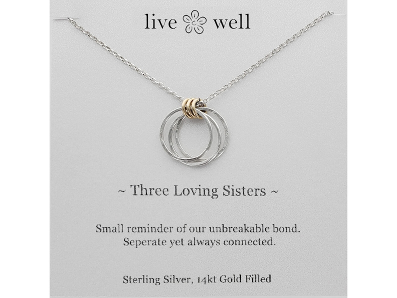 K&ouml;nnte beinhalten: Halskette aus Sterlingsilber mit drei ineinandergreifenden Ringen, akzentuiert mit goldgef&uuml;llten Details. Die Kette h&auml;ngt an einer zarten Kette und wird auf einer Karte mit dem Text "live well" und "Three Loving Sisters" pr&auml;sentiert.
