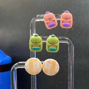 May include: A display of colorful earrings. The earrings include pairs shaped like small backpacks in pink, green, and yellow, and round earrings with a patterned design. The earrings are displayed on a clear acrylic stand.