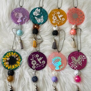 May include: A collection of round car air fresheners with various designs. Each air freshener has a different color and features images of butterflies, hummingbirds, and floral patterns. They are adorned with beads and tassels, hanging from a chain.