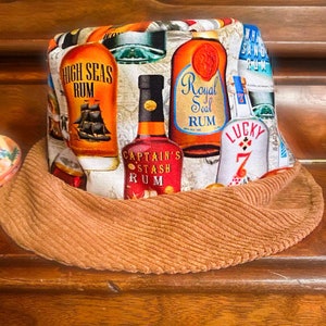 May include: A brown corduroy bucket hat with a colorful print of various rum bottles. The bottles feature labels with the text "High Seas Rum", "Captain's Stash Rum", "Royal Seal Rum", and "Lucky 7".