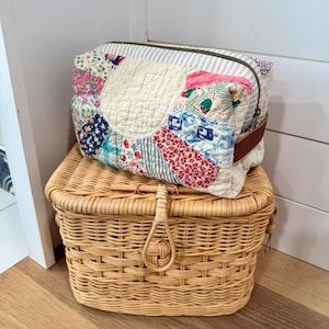 May include: A patchwork fabric cosmetic bag with a brown leather strap sits atop a woven wicker basket. The bag features a variety of floral and patterned fabrics in shades of blue, red, and pink. The basket is light brown and has a handle.