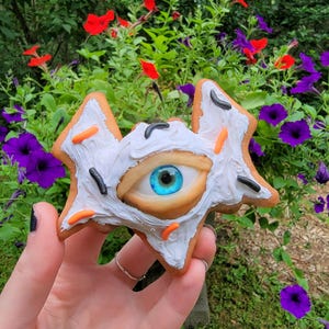 May include: A star-shaped cookie decorated with white frosting, orange and black sprinkles, and a realistic eye with a blue iris. The cookie is held in a hand against a backdrop of colorful flowers.