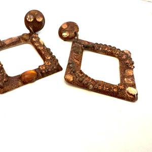 May include: A pair of dangling earrings featuring a square and a diamond-shaped frame. The frames are finished in a textured, metallic copper-brown tone and are embellished with various small, round, amber-colored beads and reflective accents.