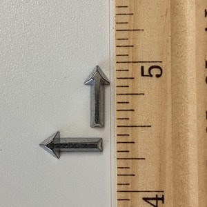 May include: Two small, silver-colored metal arrow charms. One arrow points up, the other points left. A wooden ruler with inch markings is in the background, showing the charms are approximately 0.5 inches in length.