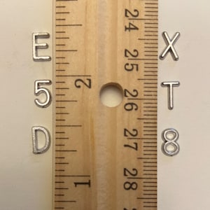 May include: A wooden ruler with inch markings next to small, silver metal letters and numbers: E, 5, D, X, T, and 8. The ruler is positioned vertically, and the letters are scattered around it.