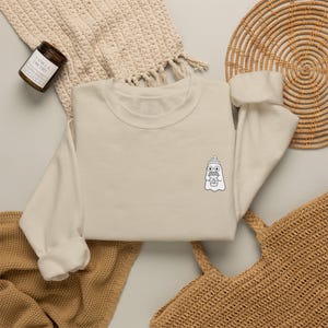 May include: A cream-colored crewneck sweatshirt featuring a small, centered black-and-white line art graphic of a ghost wearing a beanie and glasses while holding a coffee cup. The sweatshirt has long sleeves and a ribbed collar, cuffs, and hem.