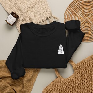 Puede incluir: Una sudadera negra de manga larga con un pequeño gráfico blanco en el pecho izquierdo. El gráfico muestra un fantasma con gorro y gafas redondas, sosteniendo una taza de café con un diseño de calabaza impreso en la parte frontal.
