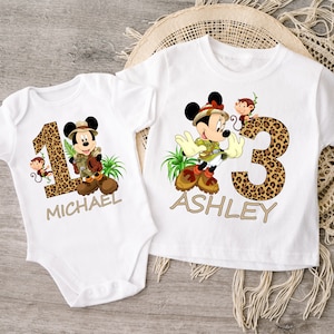 May include: Two white t-shirts with a safari theme. One shirt has a number one with a Mickey Mouse character wearing a safari hat and holding a map. The shirt says "MICHAEL". The other shirt has a number three with a Minnie Mouse character wearing a safari hat and holding a map. The shirt says "ASHLEY".