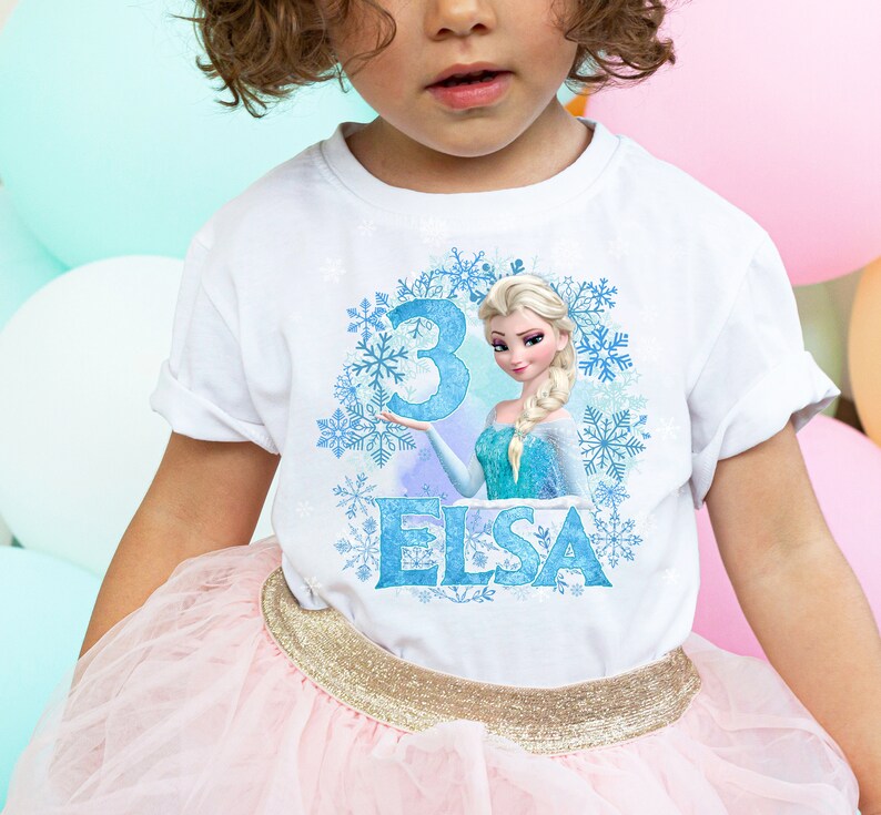 May include: White t-shirt with a blue and white graphic of Elsa from Frozen. The graphic includes a blue number 3 and the word "Elsa".