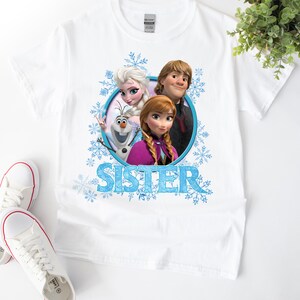 May include: White t-shirt with a graphic of Elsa, Anna, and Olaf from the movie Frozen. The graphic is surrounded by snowflakes and the word "Sister" is printed below the graphic.