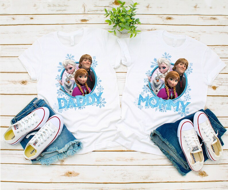 May include: Two white t-shirts with a Frozen theme. The left shirt says "Daddy" and the right shirt says "Mommy". Both shirts feature a graphic of Anna, Elsa, Olaf, and Kristoff.