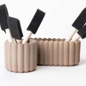 May include: Two beige, fluted containers holding black foam brushes with wooden handles. The containers are cylindrical and rectangular, and sit on a white surface. The brushes are arranged at different angles.