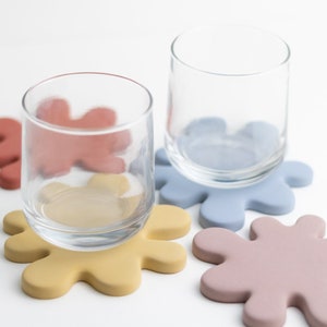 May include: Two clear glass cups on colorful silicone coasters. The coasters are shaped like irregular blobs in shades of pink, blue, and yellow.