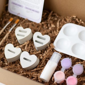 May include: A craft kit for painting heart-shaped wooden ornaments. The kit includes four unfinished wooden hearts, a small bottle of white paint, six paint pots in various shades of pink and purple, a small tray with six wells, and two paintbrushes.