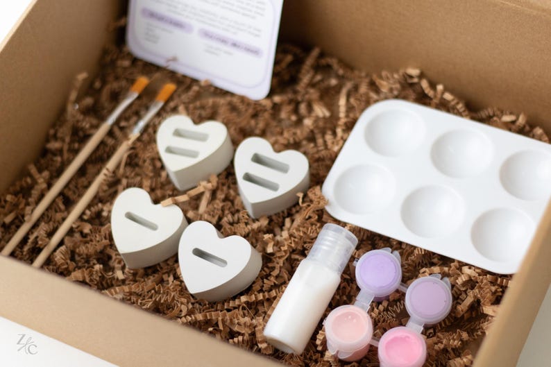 May include: A craft kit for painting heart-shaped wooden ornaments. The kit includes four unfinished wooden hearts, a small bottle of white paint, six paint pots in various shades of pink and purple, a small tray with six wells, and two paintbrushes.
