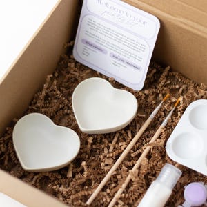 May include: A craft kit for painting ceramic heart-shaped dishes. The kit includes two white heart-shaped dishes, two paintbrushes, a paint palette, a bottle of paint, and a small bag of purple paint dots.