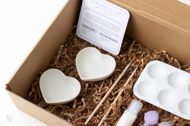 May include: A craft kit for painting ceramic heart-shaped dishes. The kit includes two white heart-shaped dishes, two paintbrushes, a paint palette, a bottle of paint, and a small bag of purple paint dots.
