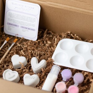 May include: A craft kit for making personalized concrete hearts. The kit includes four small white heart-shaped molds, two paintbrushes, a small bottle of sealant, six small paint pots, and a white tray with six depressions for holding paint.