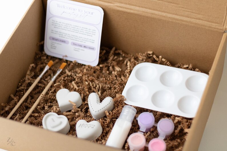 May include: A craft kit for making personalized concrete hearts. The kit includes four small white heart-shaped molds, two paintbrushes, a small bottle of sealant, six small paint pots, and a white tray with six depressions for holding paint.