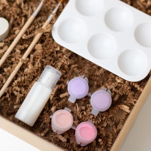 May include: A craft kit with a white heart-shaped mold, two wooden paintbrushes, a clear bottle of sealant, a white paint palette with six wells, and four small paint pots with pink and purple paint.