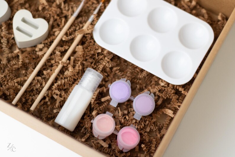May include: A craft kit with a white heart-shaped mold, two wooden paintbrushes, a clear bottle of sealant, a white paint palette with six wells, and four small paint pots with pink and purple paint.
