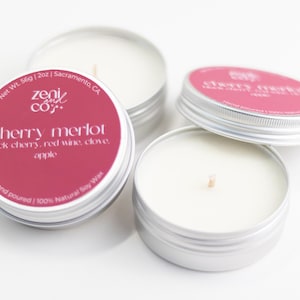 May include: Four small, silver-colored tin candles with white wax and wooden wicks. The labels are a deep red with the words "cherry merlot" and other text. The candles are hand-poured with 100% natural soy wax.