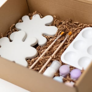 May include: A cardboard box filled with a paint-your-own pottery kit. The kit includes two white ceramic pieces shaped like abstract blobs, two paintbrushes, a white paint palette with six wells, and small bottles of paint.