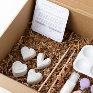 May include: A cardboard box filled with craft supplies for painting ceramic hearts. The box contains four white ceramic hearts, two paintbrushes, a small container of white paint, a small container of purple paint, and a white paint palette with six wells.